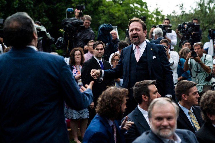 A man in a suit yells toward another man. They are surrounded by press and photographers with cameras.