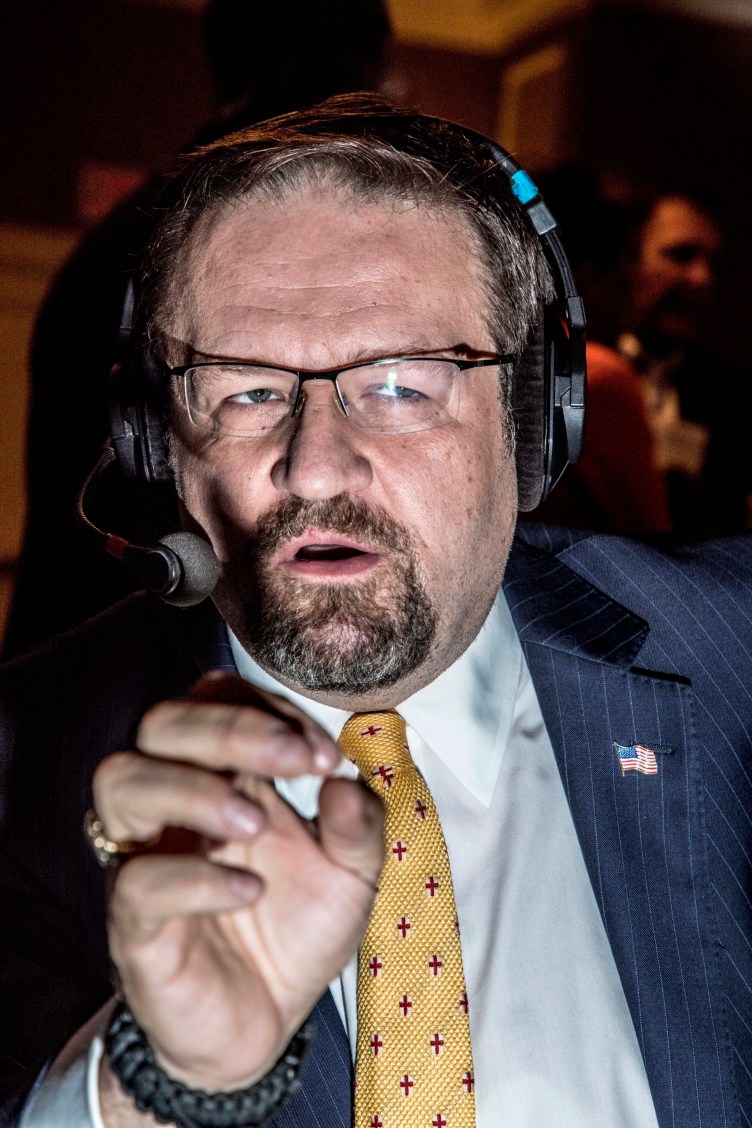 A close-up image of a man in glasses with a beard. He is wearing a suit and tie, headphones and an American flag pin. His hand is gesturing as he speaks.